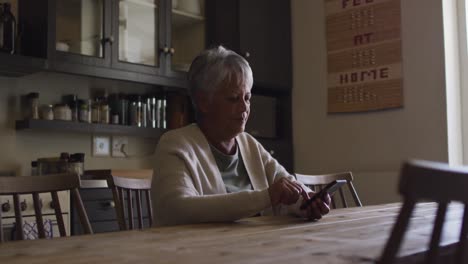 Senior-mixed-race-woman-texting-on-smartphone-sitting-in-kitchen