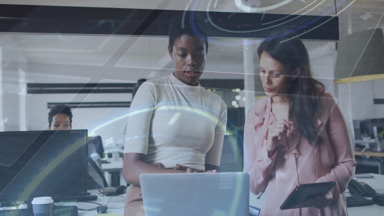 Two women collaborating over laptop in business office, with faint data charts floating overhead
