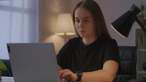 Joven-Escritora-O-Periodista-Está-Escribiendo-Texto-En-Una-Computadora-Portátil-Trabajando-En-Casa-Por-La-Noche-Sentada-En-La-Mesa-En-La-Habitación-Del-Apartamento-Retrato-De-Una-Mujer-Independiente-En-El-Trabajo