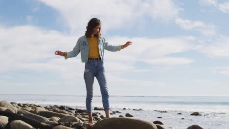 happy mixed race woman walking barefoot on rocky beach by the sea in the sun