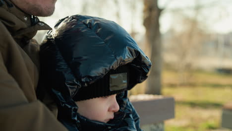 close-up of a father and son sitting closely together on a bench, both looking focused, the son wears a black jacket and the father a brown jacket, with a blurred background of nature