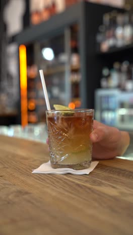 a dark and stormy cocktail served in an intricately designed glass with ice and garnished with a lime wedge. the drink rests atop a wooden bar counter, with a blurred bar background.