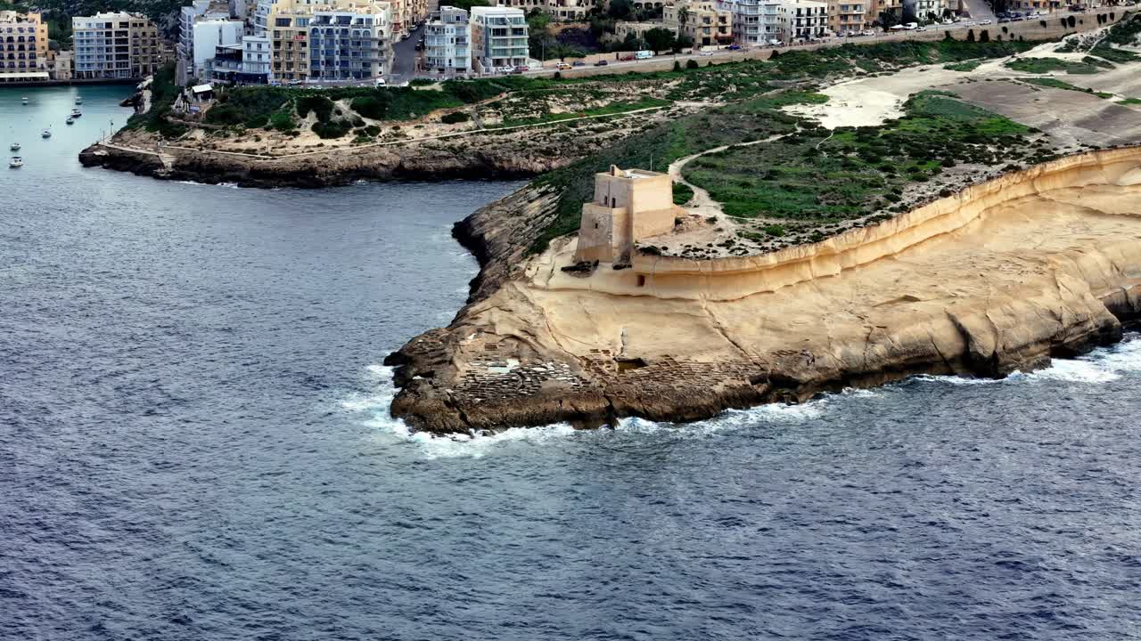 Vista panoramica aerea della Torre di Xlendi che si erge sulle frastagliate scogliere calcaree dell'isola di Gozo a Malta durante una luminosa giornata estiva di sole, con acque turchesi del Mediterraneo e panorama della città costiera