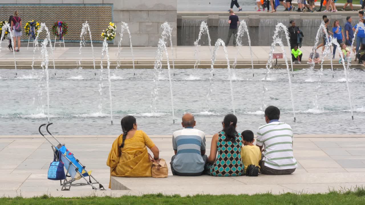 Indian family sits by National World War II Memorial square and flowing water fountain, Washington, D.C., behind view