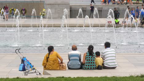 indian family sits by national world war ii memorial square and flowing water fountain, washington, d.c., behind view
