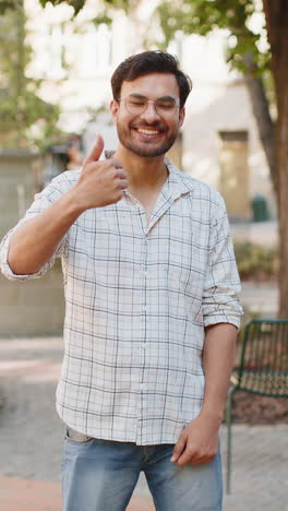 happy young indian man looking approvingly at camera showing thumbs up like gesture on city street