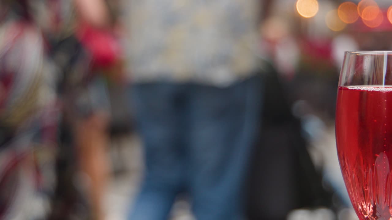 Camera focuses from blurred street scene to sharp red cocktail glass, evening outdoor café ambiance