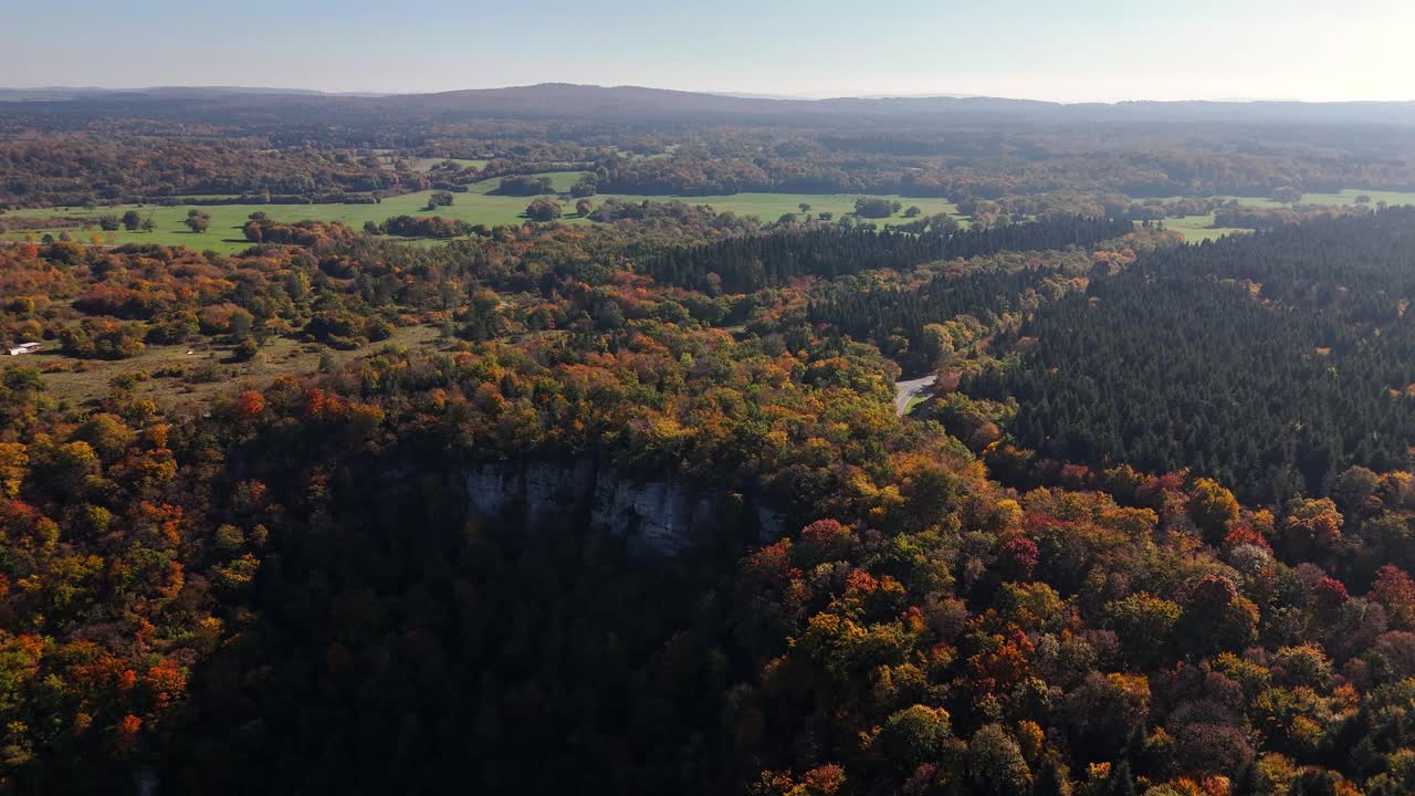 Aerial view of Cirque du Fer à Cheval, France revealing dense autumn forest, rugged cliff edges, winding road and expansive green valley stretching toward distant hills beneath a bright and clear sky