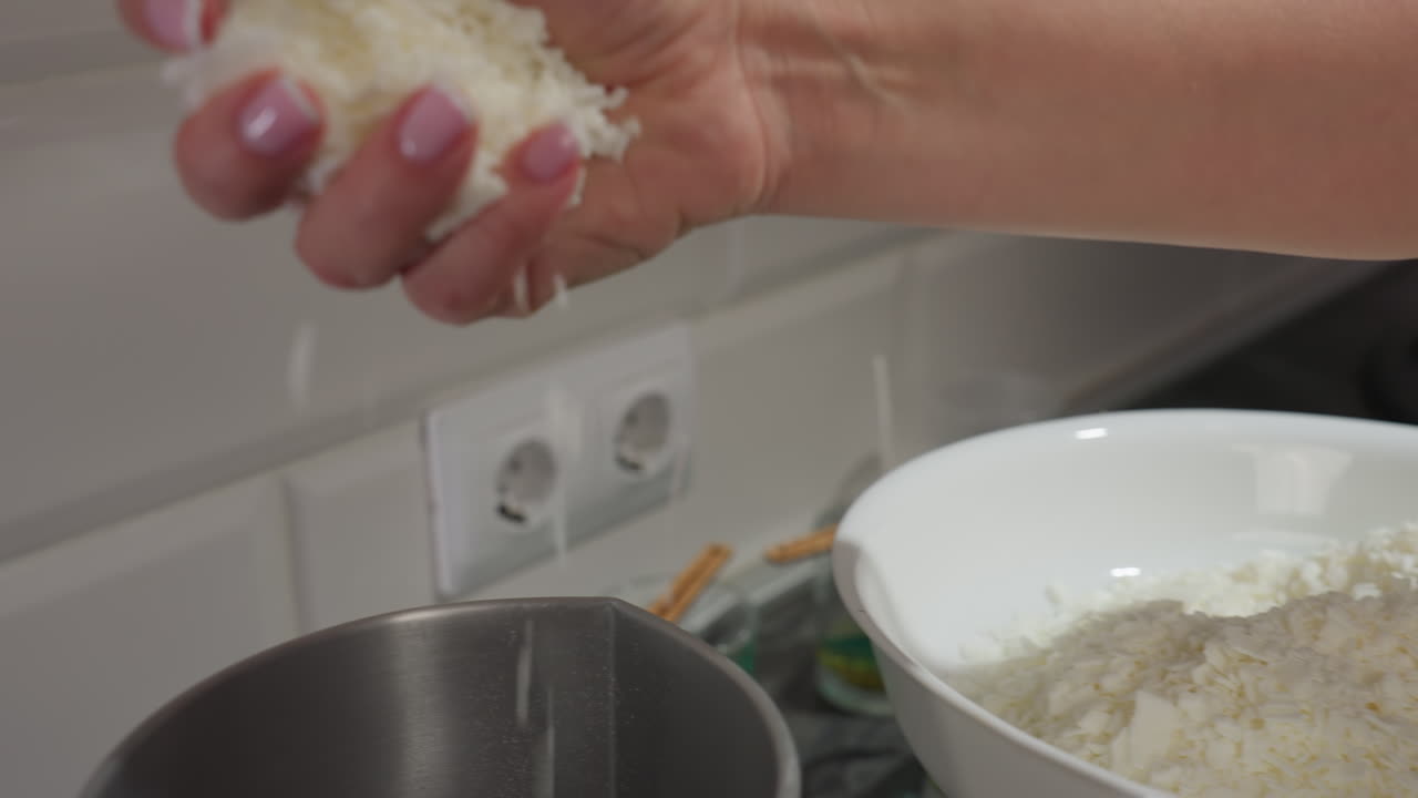 Elegant woman with manicured hands scooping white baking mixture from bowl into mixer on kitchen countertop with visible green candle jars in background showing cooking preparation in progress