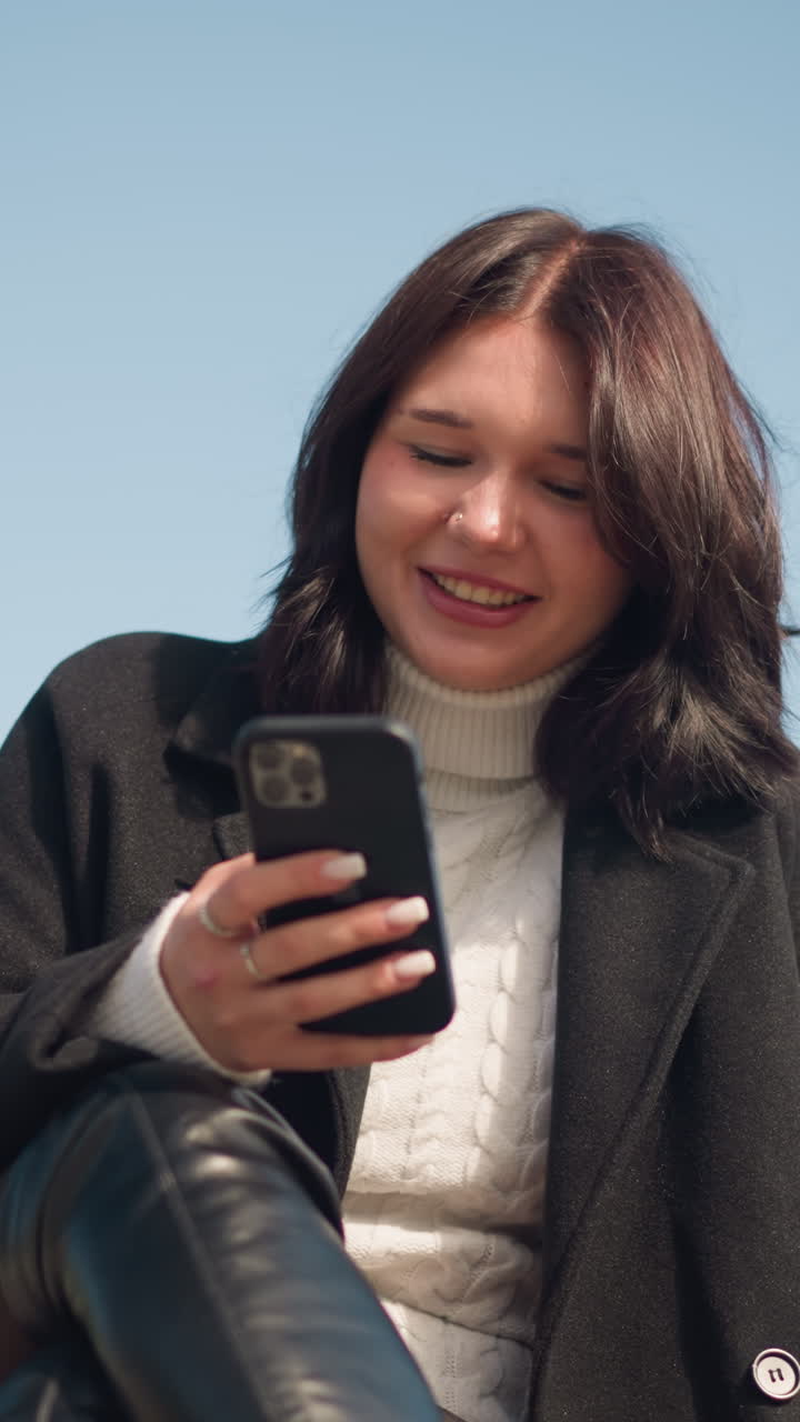 dama profesional sonriendo mientras se centra en el teléfono inteligente en un entorno de parque, con abrigo negro y suéter blanco, con las piernas cruzadas, disfrutando de una atmósfera al aire libre serena bajo un cielo azul y una vegetación exuberante