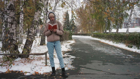 light skin woman standing on snowy park path wearing winter clothing while reading book under autumn trees, focused on pages as leaves and snow surround peaceful quiet outdoor setting in cold weather