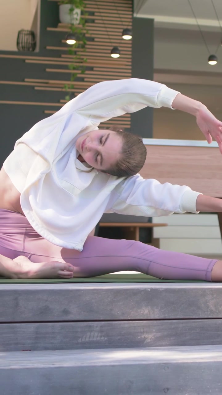 Vertical video: Extending right leg sweatshirt woman holding foot while partner twisting on mats