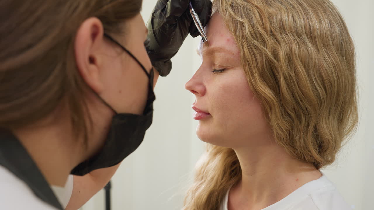 Beautician wearing black gloves precisely applies cream to upper eyebrow of client with closed eyes. Client with wavy blonde hair remains calm in well-lit beauty salon during treatment session