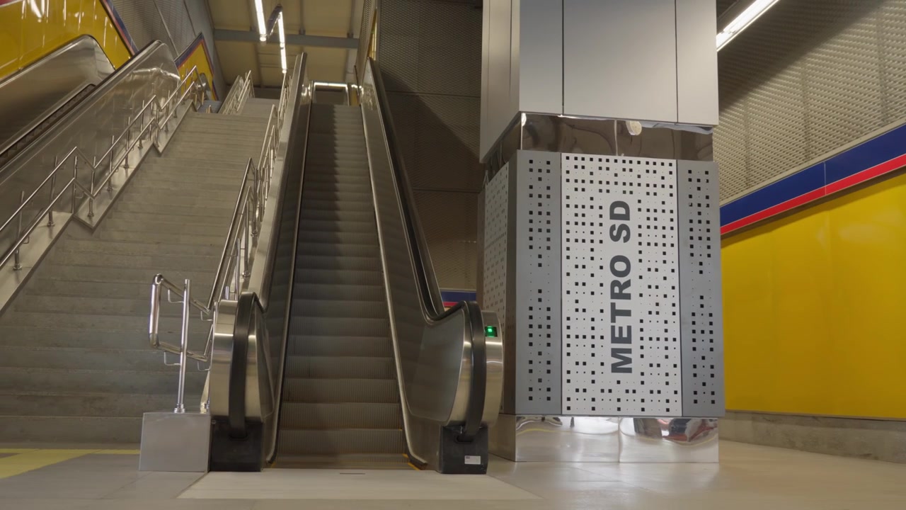Un plano secuencia que pasa junto a un pilar con la marca Metro SD hacia escaleras mecánicas ascendentes y escaleras de hormigón dentro de una moderna estación de metro.