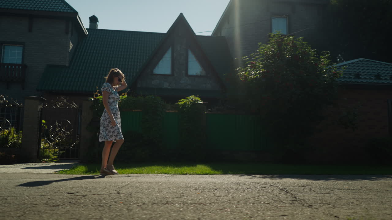 Elegant woman in floral dress walks past suburban home while putting on sunglasses as sunbeam casts gentle light on her face and body, creating serene and stylish moment in quiet neighborhood
