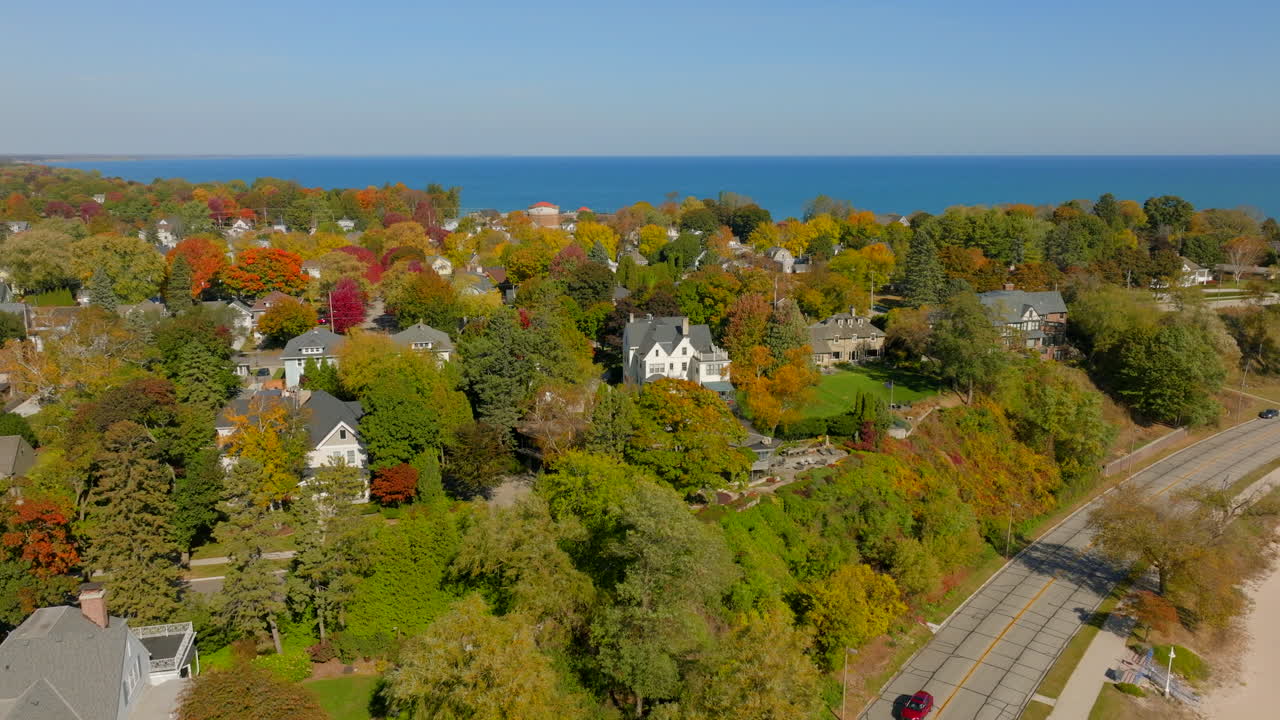 Drone aerial descending over Sheboygan, Wisconsin neighborhood homes and autumn trees with Lake Michigan beyond, moving down toward the beach road and sidewalk below under a clear blue sky