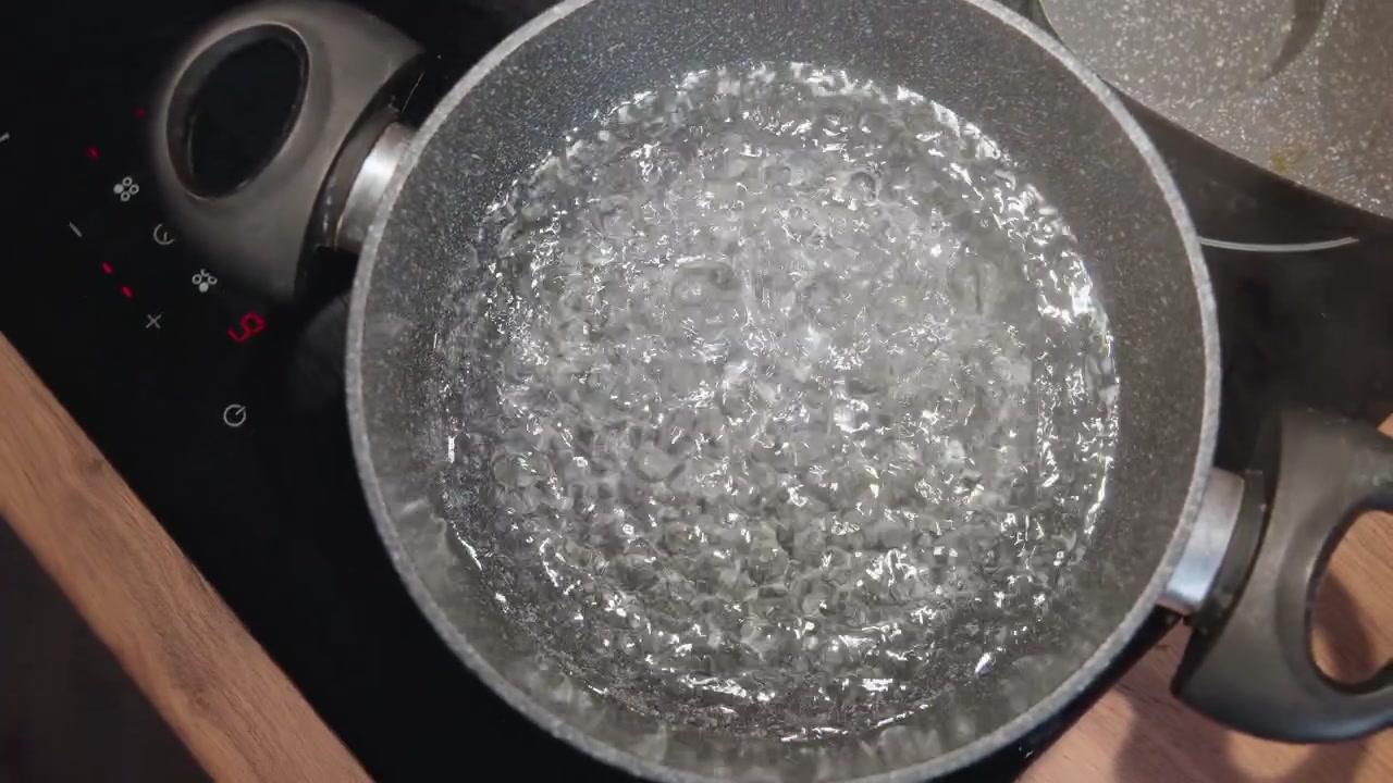 Close overhead view of metal pot on stovetop filled with rapidly boiling water, showing strong bubbling, swirling motion, shimmering reflections, splashing droplets, and visible steam rising in warm