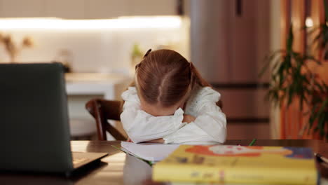 una niña parece cansada y abrumada mientras hace su tarea escolar en la cocina de una casa.