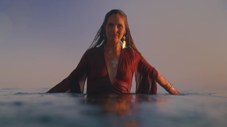 A woman stands waist deep in the sea wearing a flowing red dress at sunset.
