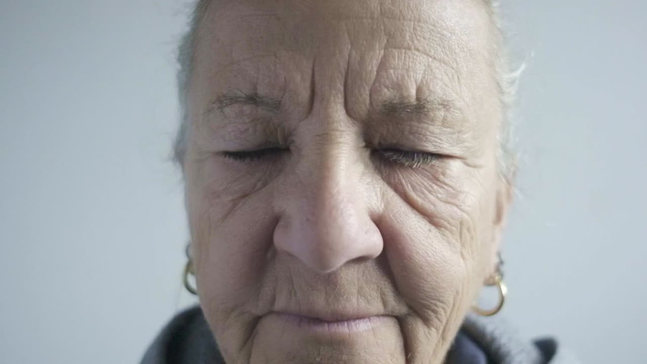 anciana mujer caucásica mira a la cámara arrugas de primer plano parpadeando ojos azules cabello blanco con pendientes y ropa deportiva, retrato facial