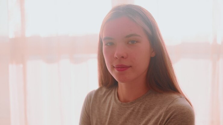 Lady seated close to window focusing on something then lifting head with confident look as natural light streams into room through curtain creating soft warm glow around her silhouette and profile