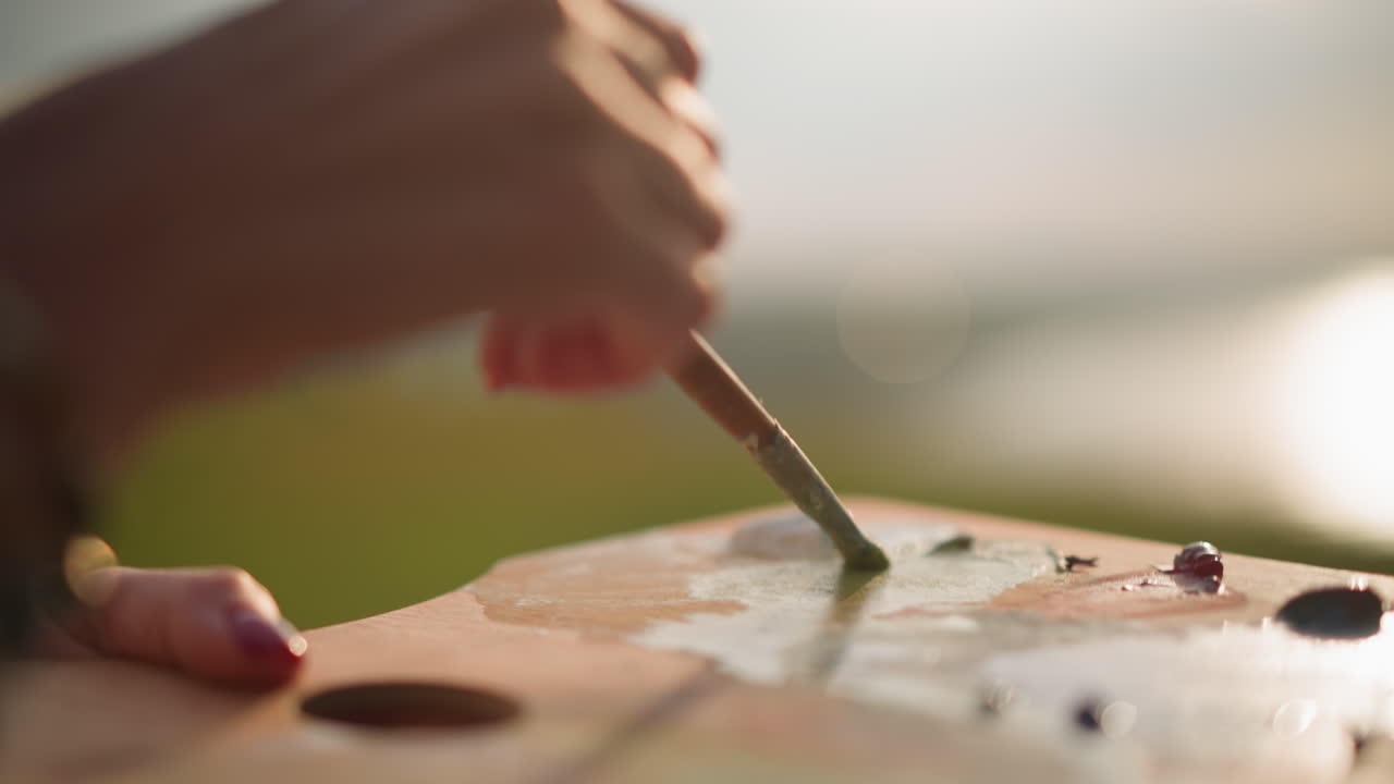 una toma de primer plano de la mano de una mujer, con uñas pintadas de rojo, mezclando cuidadosamente los colores en una paleta de madera con un pincel bajo la luz solar natural. la escena captura el momento preciso de la mezcla de colores