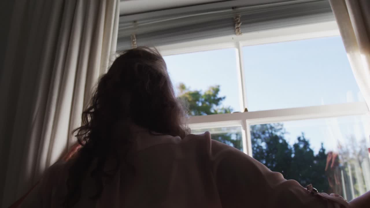 Senior caucasian woman drawing curtains in sunny room and looking out of window