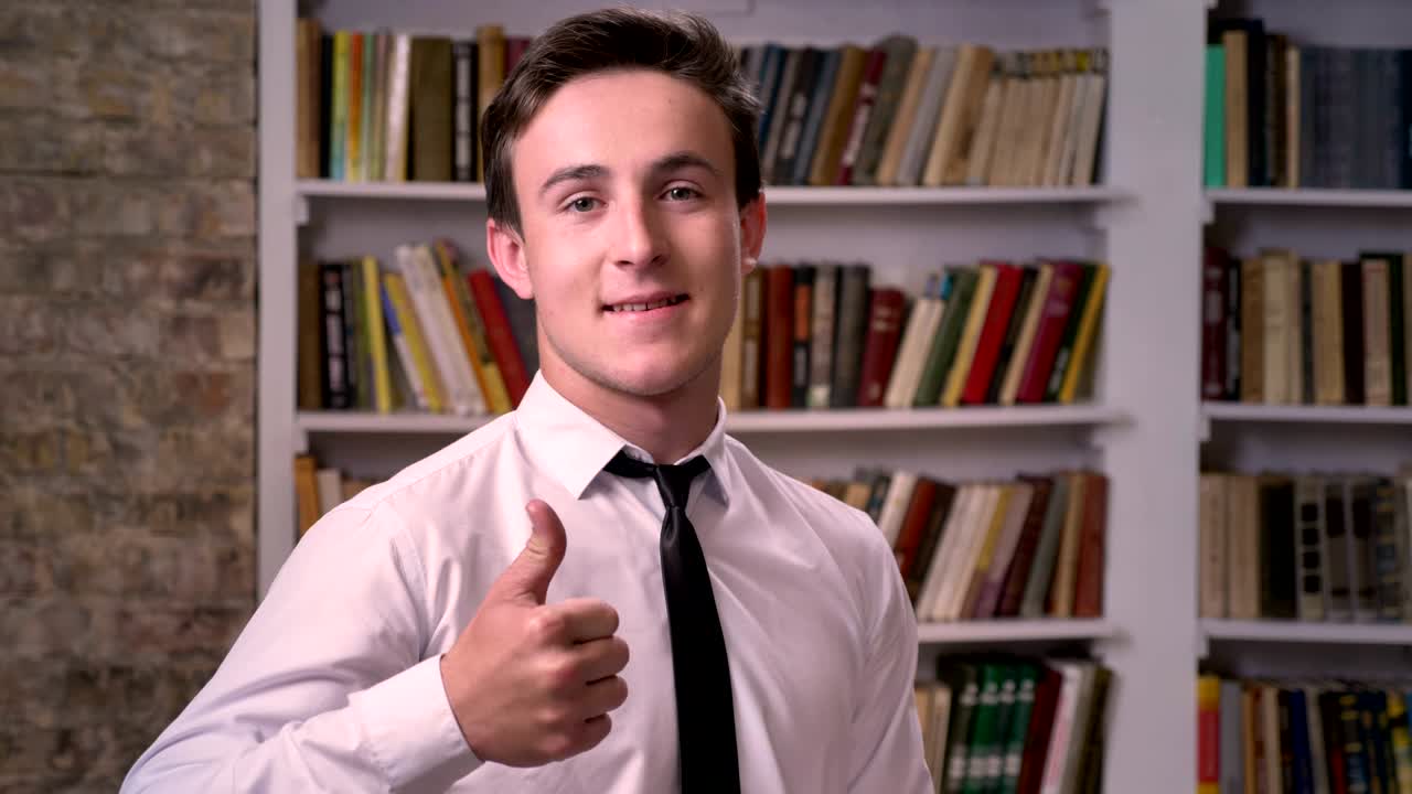 매력적인 젊은 사업가가 돌아서서 카메라를 바라보며, 표지판, 제스처 개념, 배경에 있는 책장을 보여줍니다.