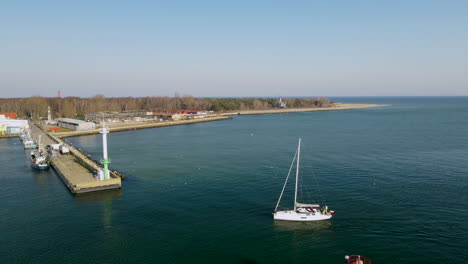 small white sailboat enters the harbor with sails rolled up, calm water of puck bay, hel, poland aerial