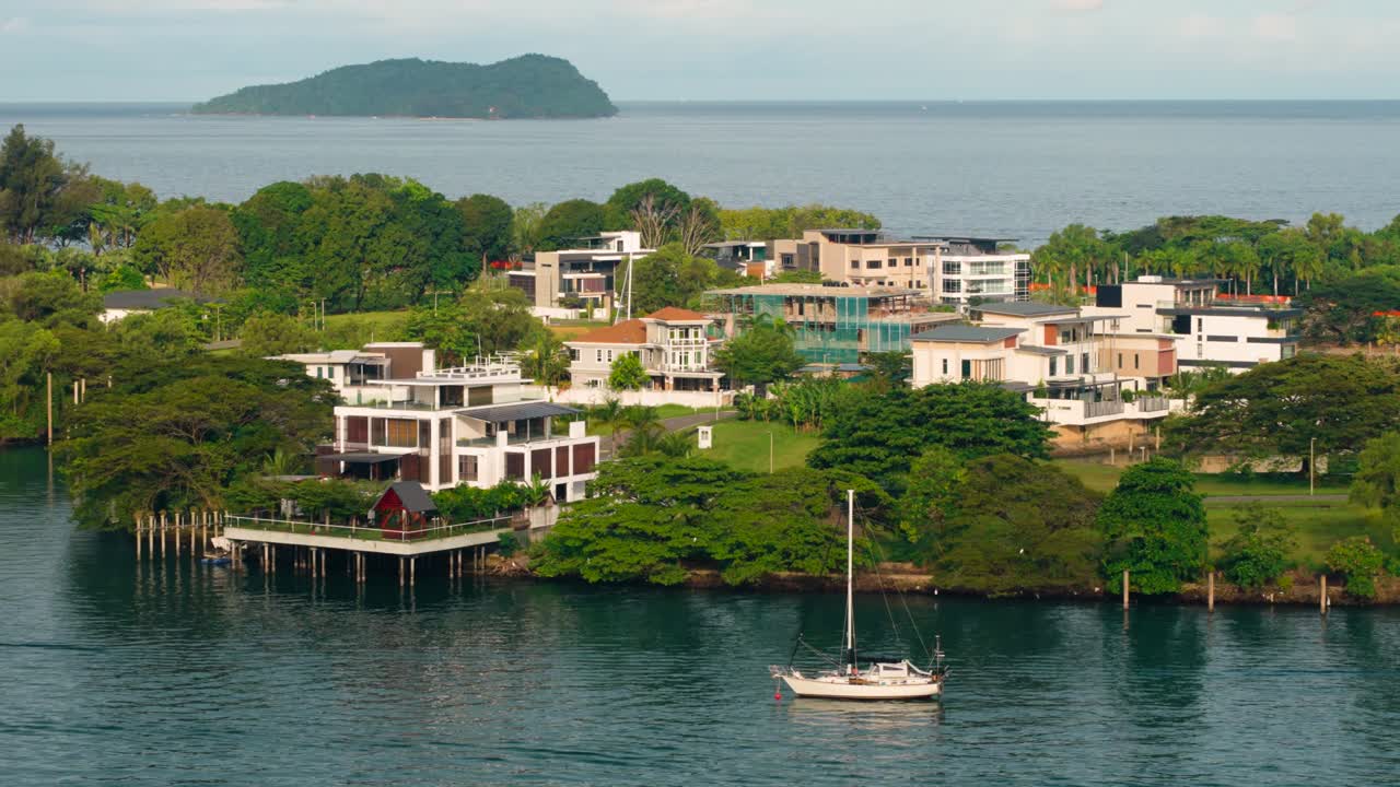 Aerial panorama of luxury Sutera residency waterfront villas in Kota Kinabalu with a white sailboat anchored in calm blue water and a tropical island on the horizon