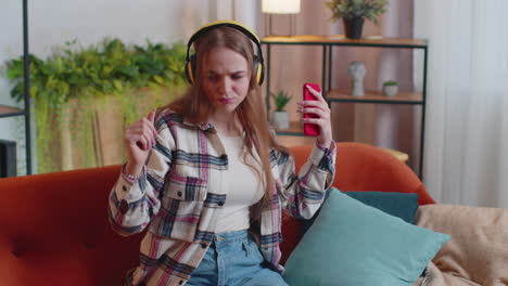 overjoyed young woman in wireless headphones dancing, singing on cozy couch in living room at home