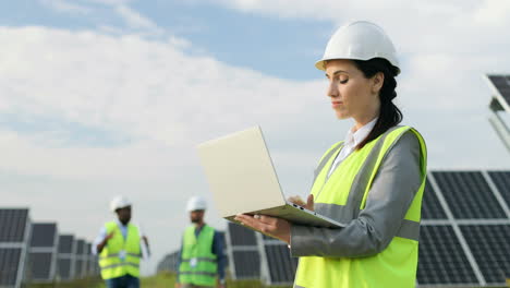 portrait of beautiful female engineer in protective helmet and uniform using laptop and looking around on solar plantation