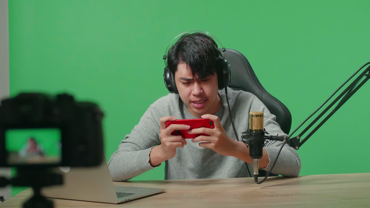 hombre asiático molesto jugador con auriculares y computadora perdiendo juego de teléfono móvil mientras graba video por cámara en el fondo de la pantalla verde