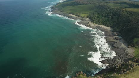 Aerial-pan-down-over-green-meadow-rocky-beach,-texture-ocean-waves