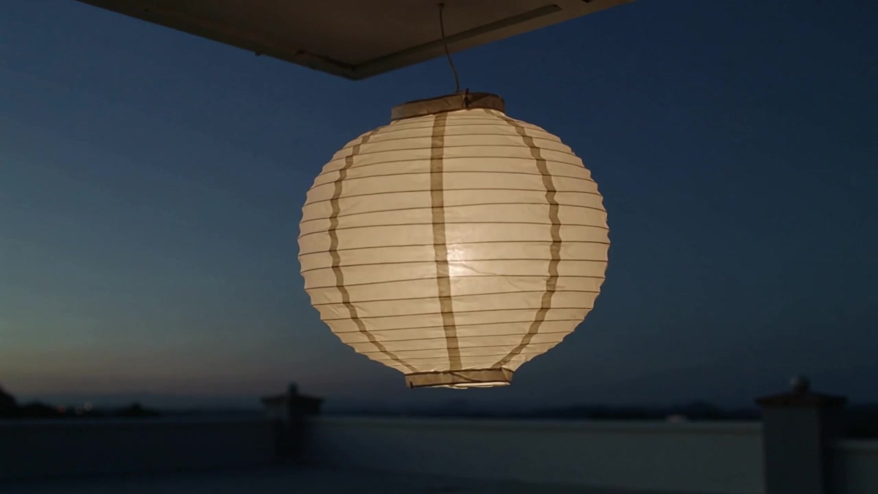 Lanterna di carta rotonda che oscilla e brilla caldamente sotto la sporgenza del tetto al crepuscolo, mossa da una leggera brezza.