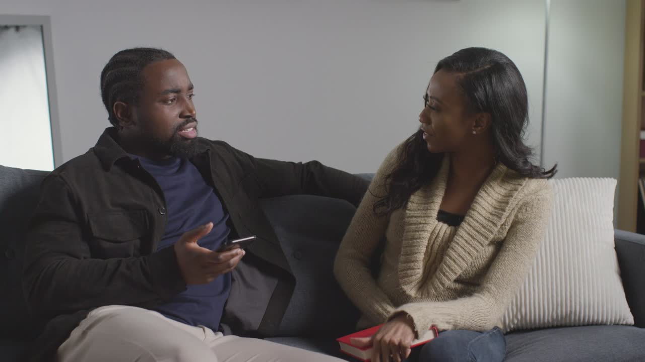 Couple At Home Sitting On Sofa In Lounge Having Argument Over What Man Is Looking At On Mobile Phone