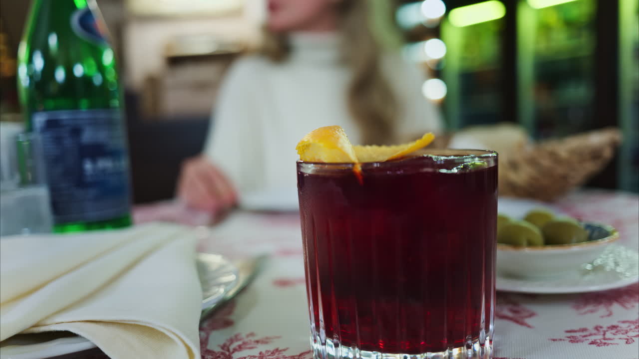 Close up of a negroni cocktail on a red and white tablecloth with a woman sitting at a restaurant on the background. Vertical