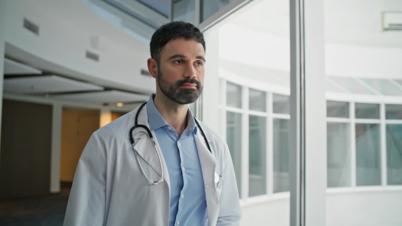 Doctor barbudo caminando por el pasillo de la clínica de cerca. Profesional concentrado pensando.
