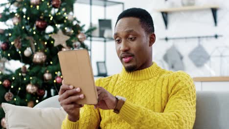 Close-Up-Portrait-Of-Happy-Man-Tapping-On-Tablet-And-Buying-Xmas-Gifts-Online-On-New-Year-Sale-Sitting-Near-Christmas-Tree-In-Room