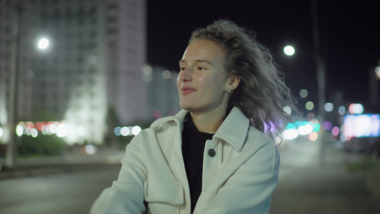 Smiling joyful girl flips hair with cheerful expression on city street at night, radiating happiness and confidence with glowing streetlights and urban blur in background