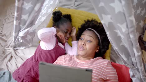 happy african american mother and daughter lying in tent and using tablet at home, slow motion