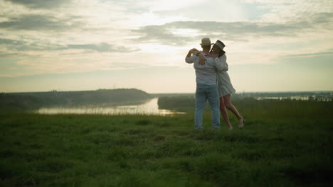 a loving man spins his wife around on a grassy hill beside a peaceful lake at sunset. the man, dressed in a white shirt, hat, and jeans, shares a tender and joyful moment with his wife