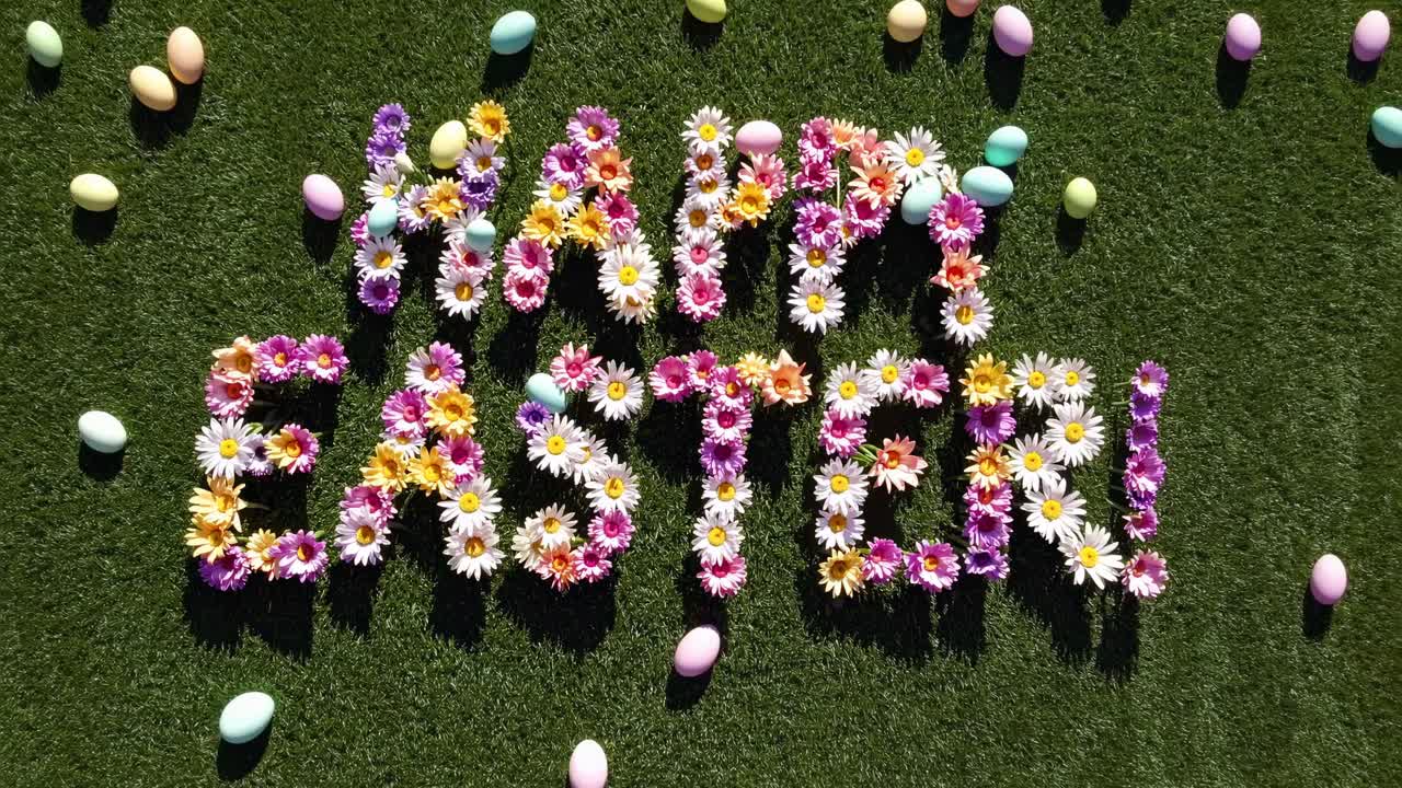 Top-down view of 'Happy Easter' spelled with flowers on grass, surrounded by colorful eggs