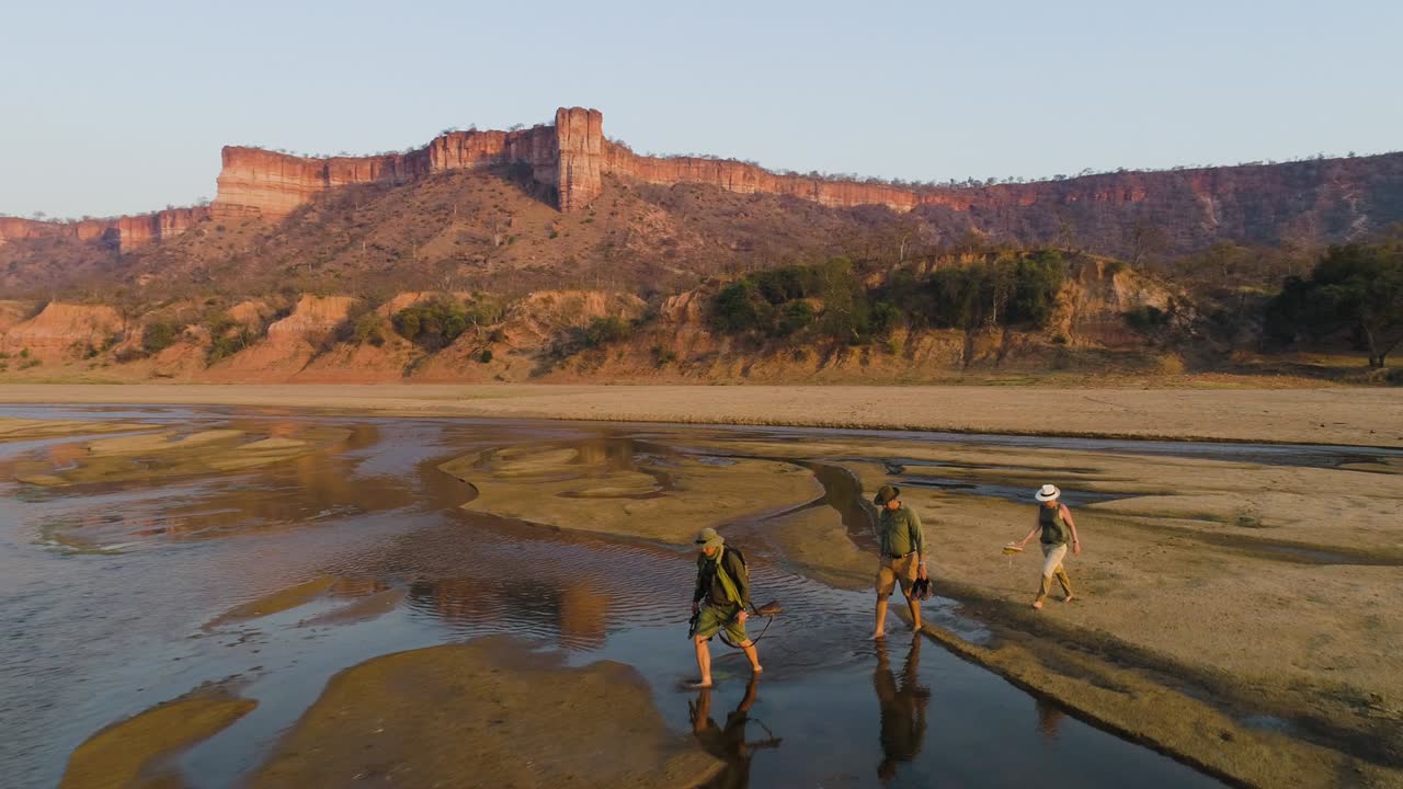vista aérea de turistas en una caminata guiada cruzando el río runde con los hermosos acantilados de piedra arenisca roja de chilojo en el fondo, parque nacional gonarezhou, zimbabue