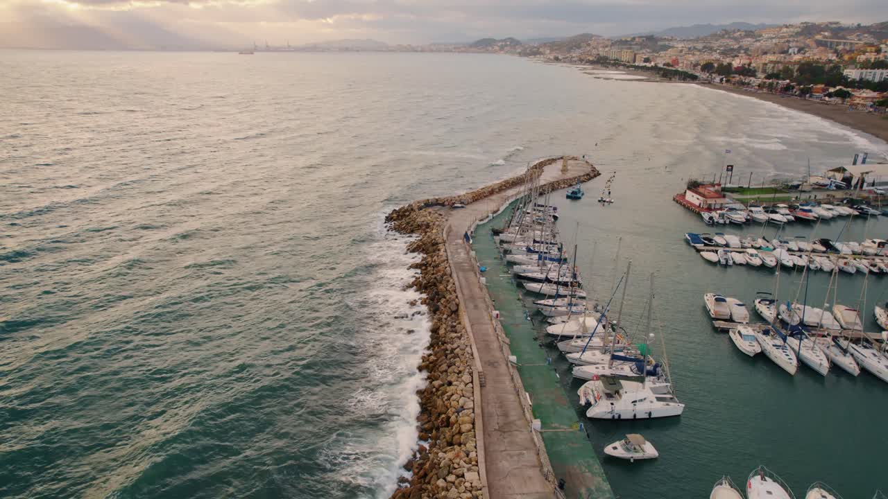 puerto marítimo con barcos y yates junto a un rompeolas rocoso y un mar con olas bajo el sol dorado al atardecer