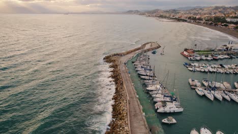 Puerto-Marítimo-Con-Barcos-Y-Yates-Junto-A-Un-Rompeolas-Rocoso-Y-Un-Mar-Con-Olas-Bajo-El-Sol-Dorado-Al-Atardecer