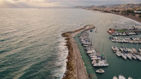 Sea-port-with-boats-and-yachts-next-to-a-rocky-breakwater-and-a-sea-with-waves-under-the-golden-sun-at-sunset