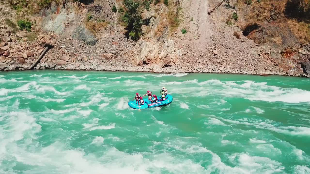Equipo de rafting remando por rápidos turquesa en el río Ganges, en un cañón de montaña cerca de Rishikesh. Toma aérea suave con seguimiento hacia adelante que sigue la balsa río abajo, pasando por terreno rocoso y zonas boscosas