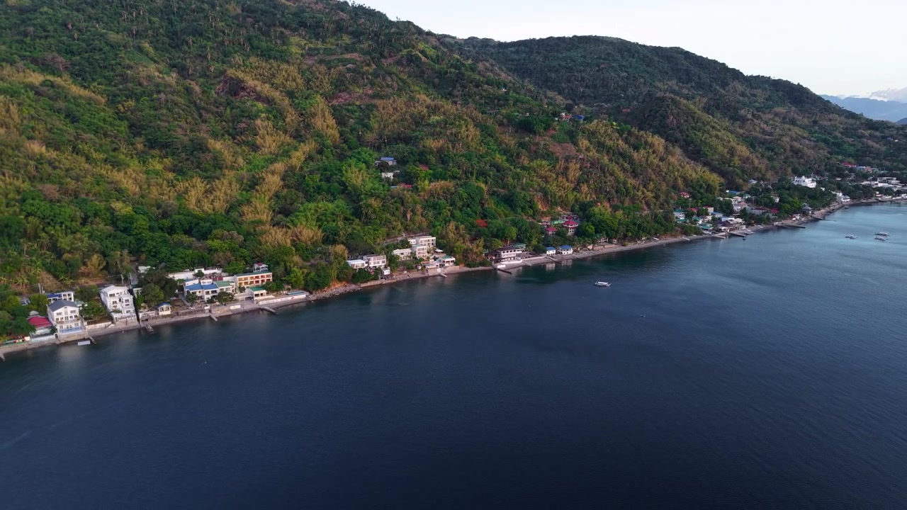 La luce dorata del sole svanisce lungo la costa di Anilao mentre un drone scivola sopra il mare tranquillo, rivelando un paesaggio costiero drammatico e un cielo colorato.