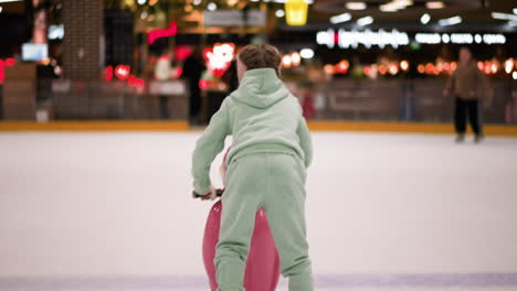 back view of a child in a mint green tracksuit and pink ice skates learning to skate using a pink training aid on an ice rink